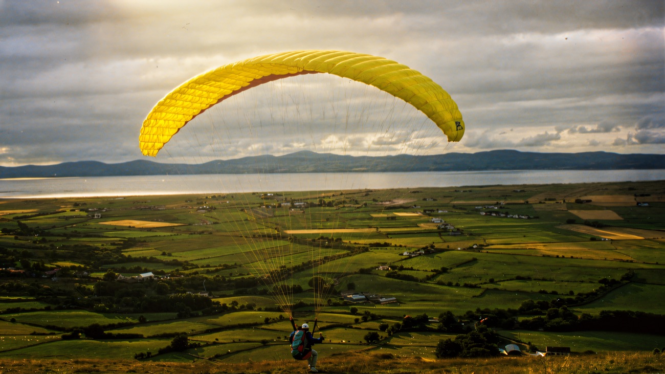 A yellow paraglider soaring above patchwork green fields, with a wide lough and blue hills on the horizon