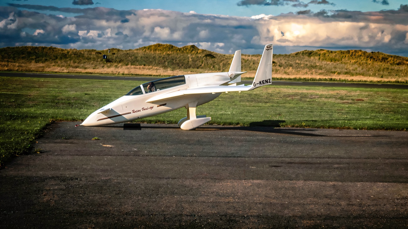 A small white aircraft parked on the tarmac beside a grass strip, with dunes rolling away behind