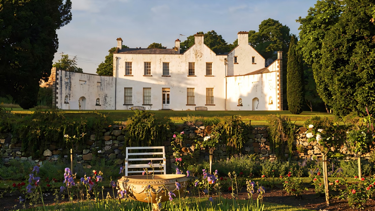 The white house at Ardress seen across the walled garden, with flower borders and a stone wall in the foreground