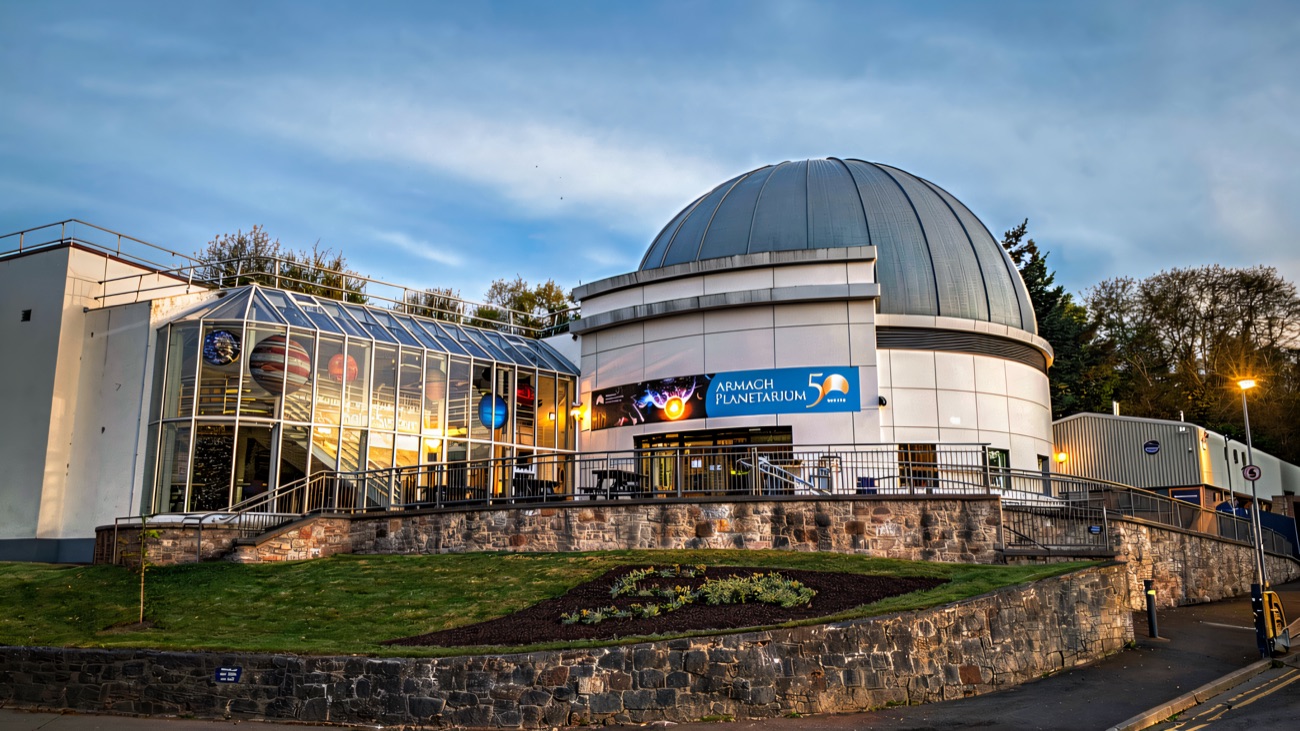 The silver planetarium dome by day beside the glass model-planets walkway under a blue sky