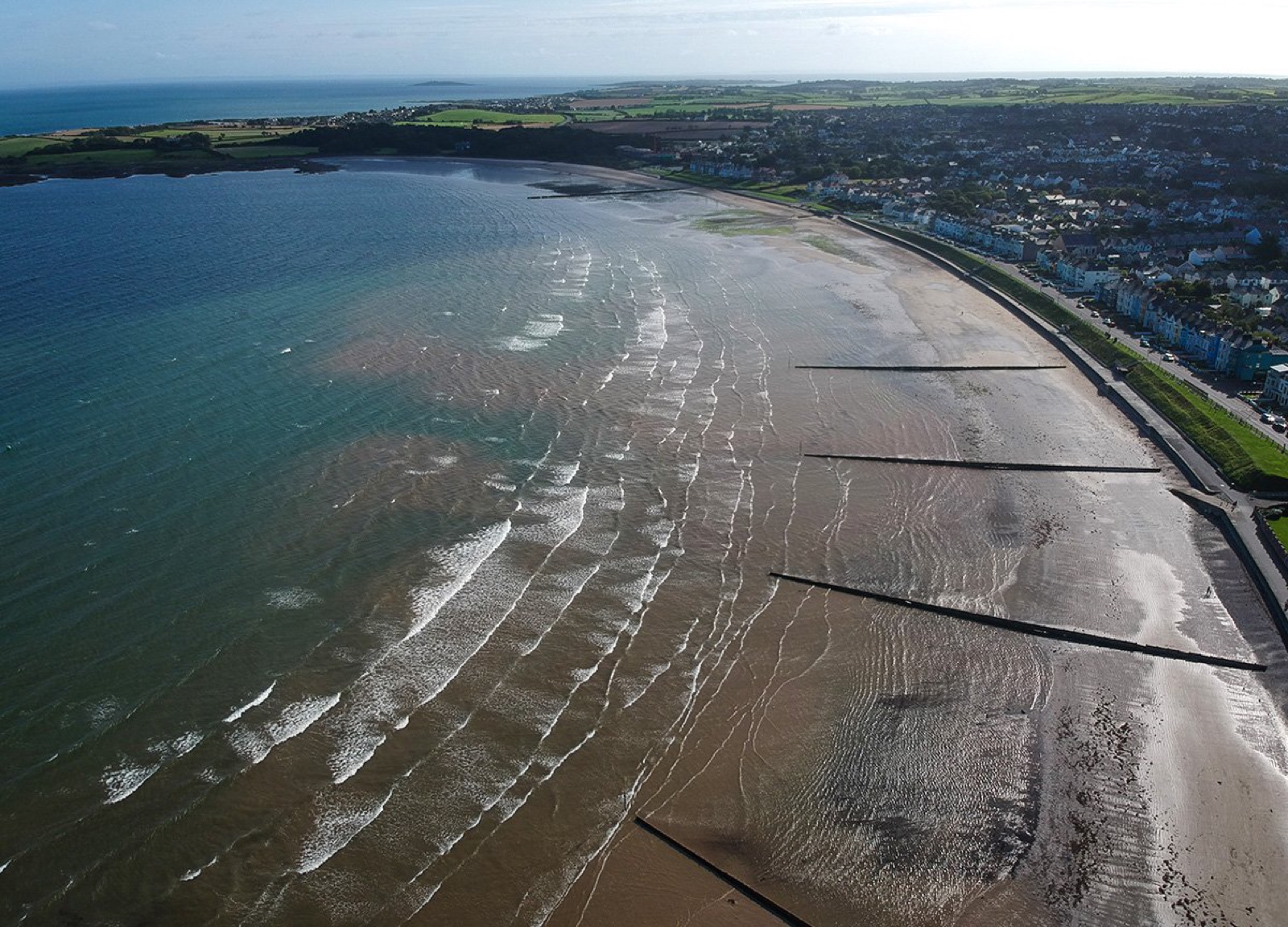 Aerial view down the long sweep of Ballyholme Beach in Bangor, waves rolling onto golden sand past timber groynes with the seafront promenade and terraced houses running along the bay