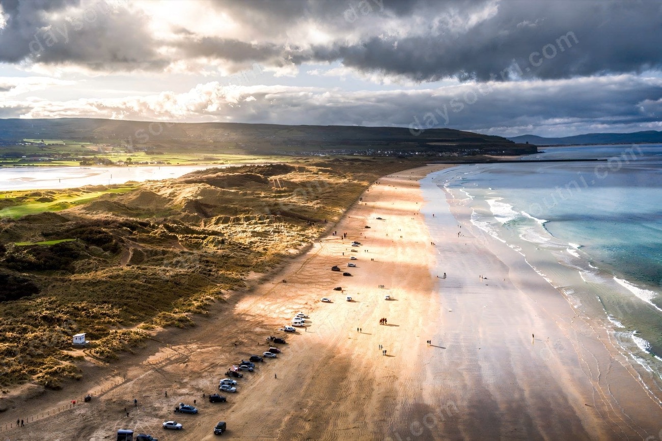 Aerial view of cars parked along the golden sand of Portstewart Strand with dunes and sunlit hills behind