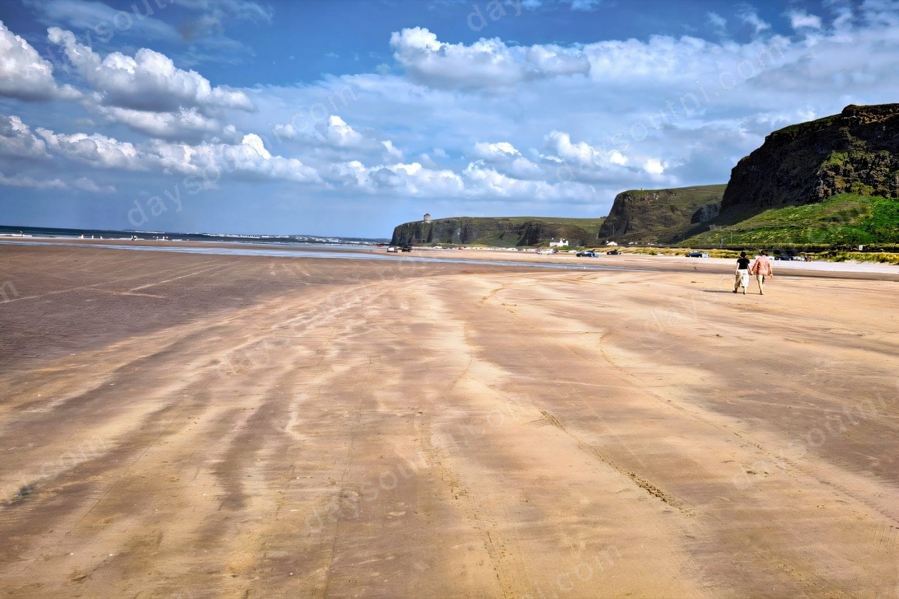 Two walkers on the wide golden sand at Benone Strand with the Binevenagh cliffs and Mussenden Temple in the distance