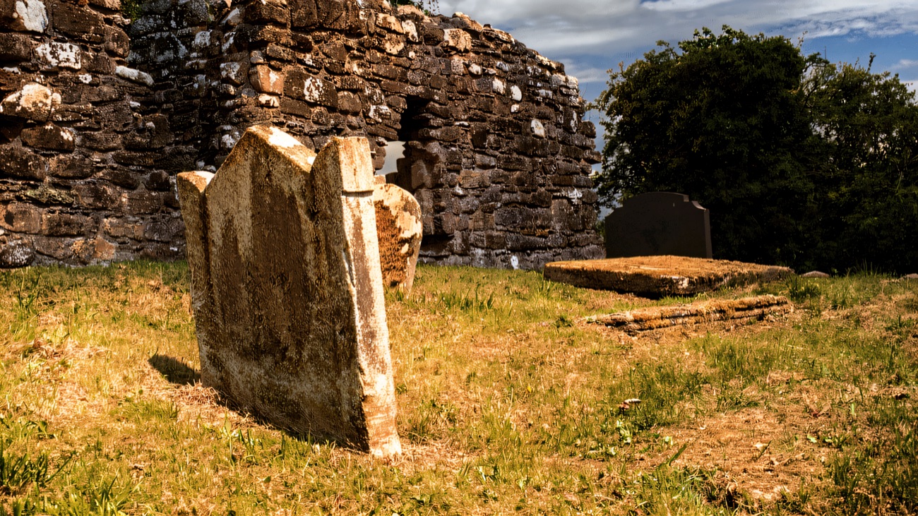 An old headstone leans in the grass before the roofless church walls on a bright day.