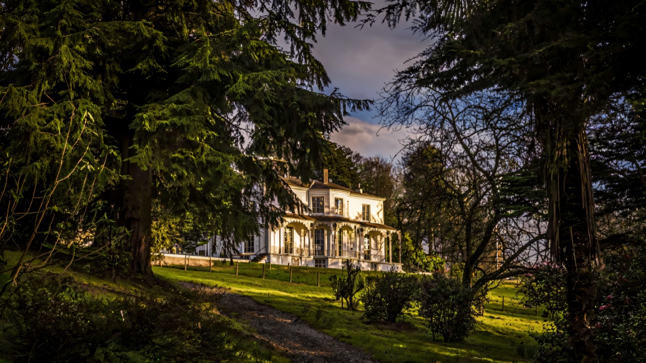 The white Georgian house at Brook Hall framed by tall trees, a gravel path leading up