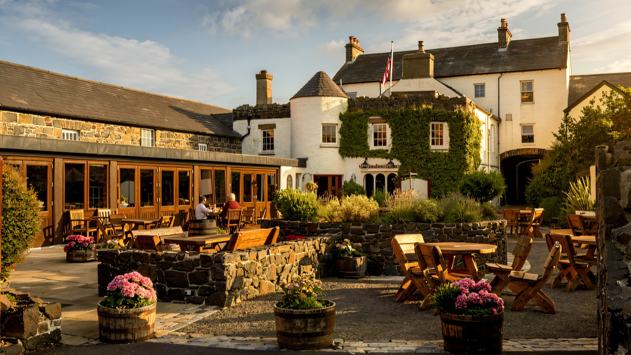 The sunny courtyard garden at the Bushmills Inn, with wooden tables, whiskey-barrel planters of pink flowers and the ivy-covered inn behind