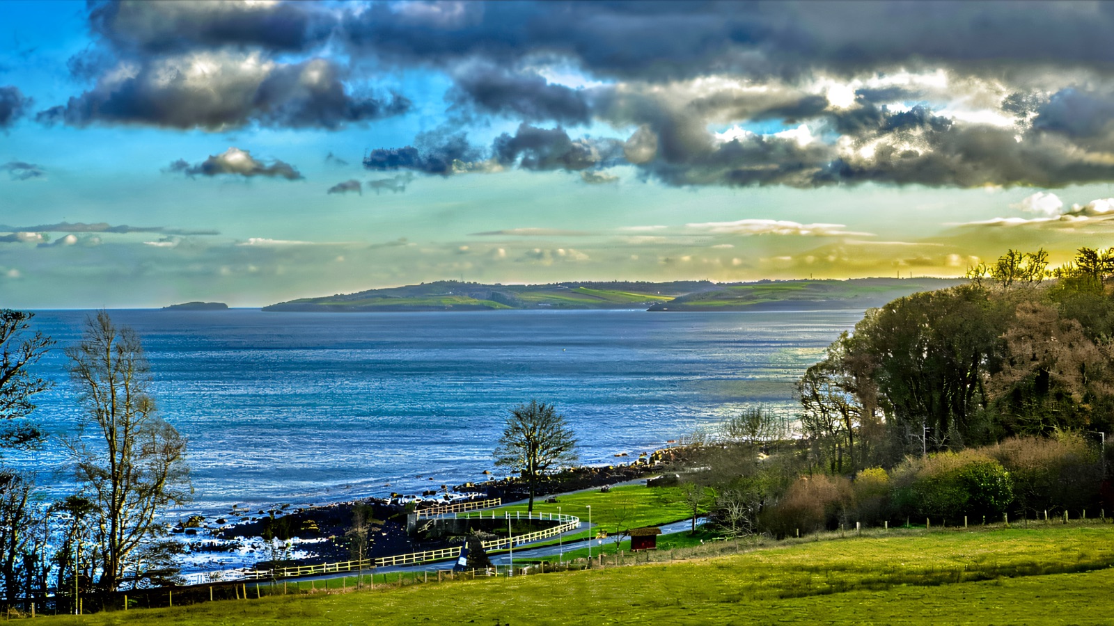 The sea and a green headland seen from Carnfunnock Country Park, with grass, a shore path and trees running down to the water