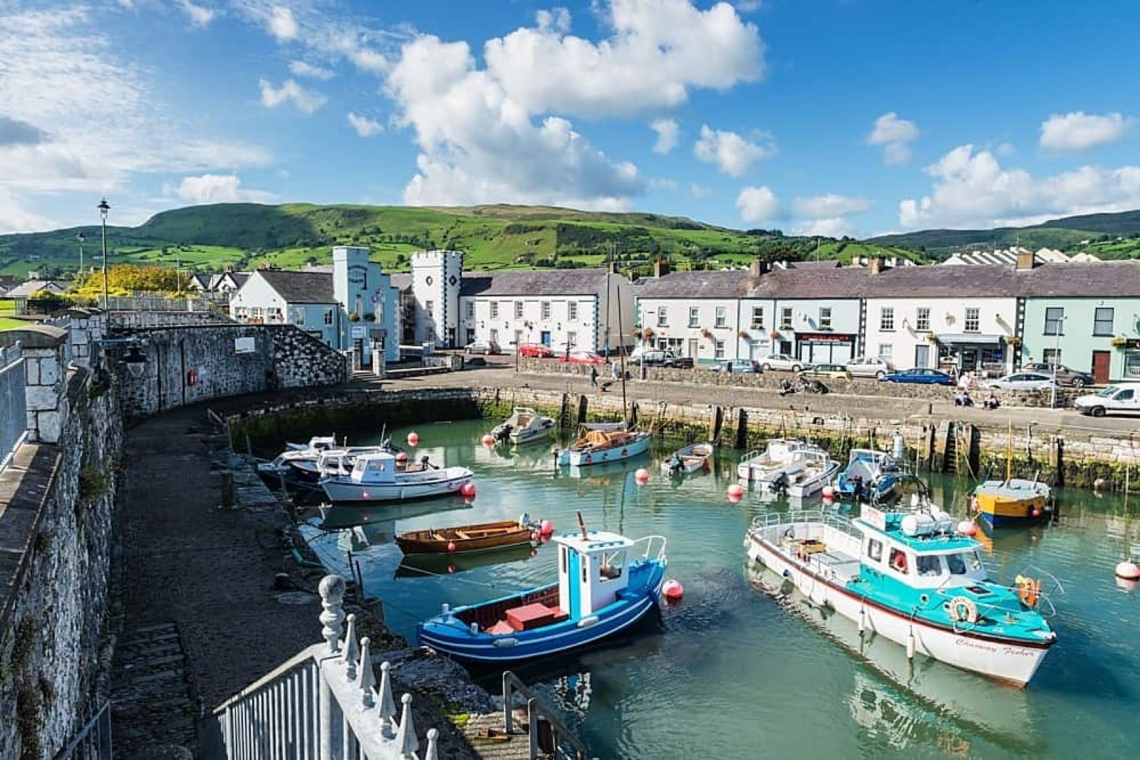 Carnlough harbour on a bright day with colourful fishing boats moored in the basin, the white houses of the village behind and green Glens hills rising above
