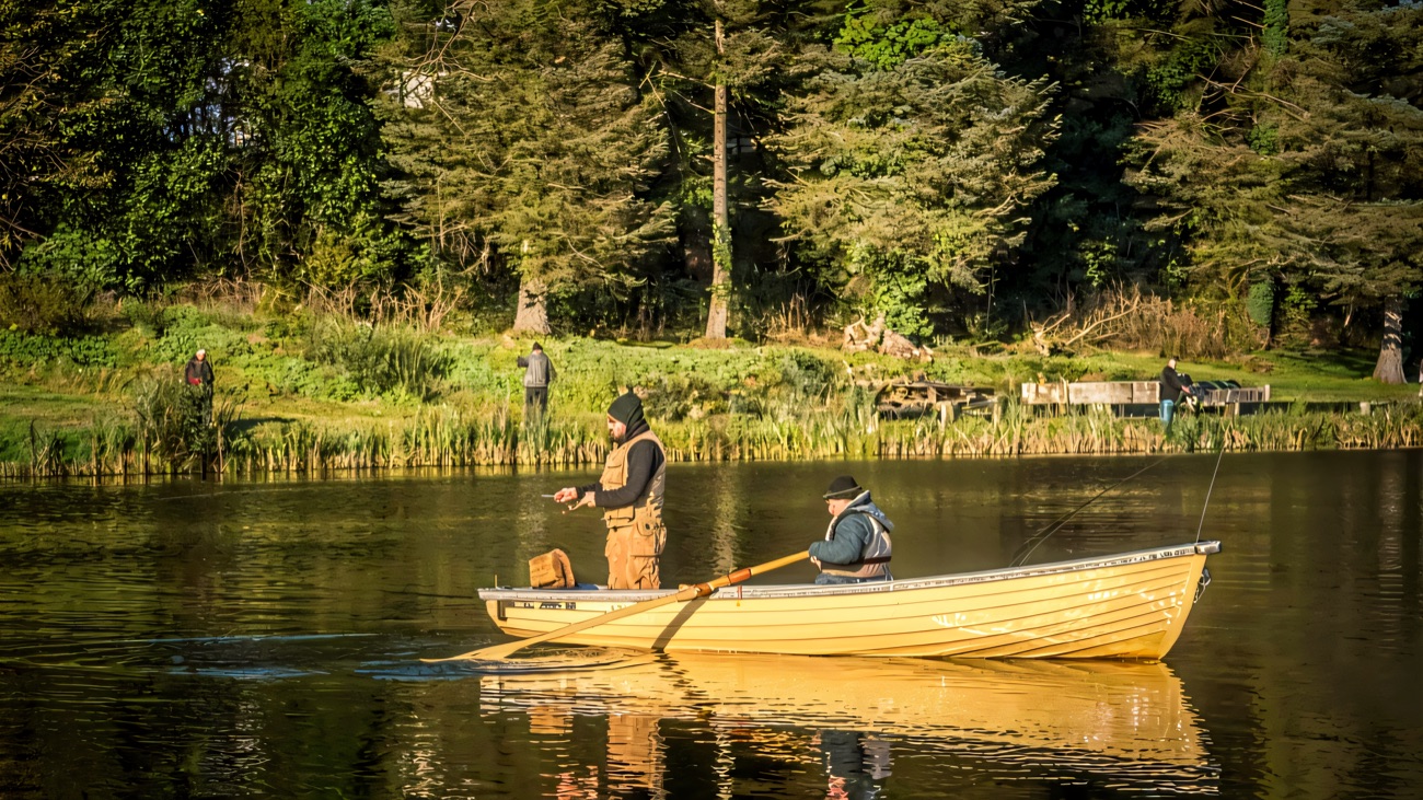 Two anglers out in a yellow rowing boat, with more fishing from the grassy bank and the jetty behind