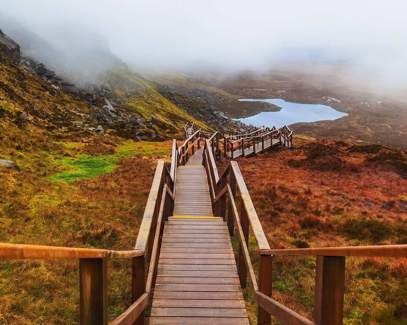 A steep wooden staircase climbs the mountainside with a small mountain lough and misty ridge beyond.