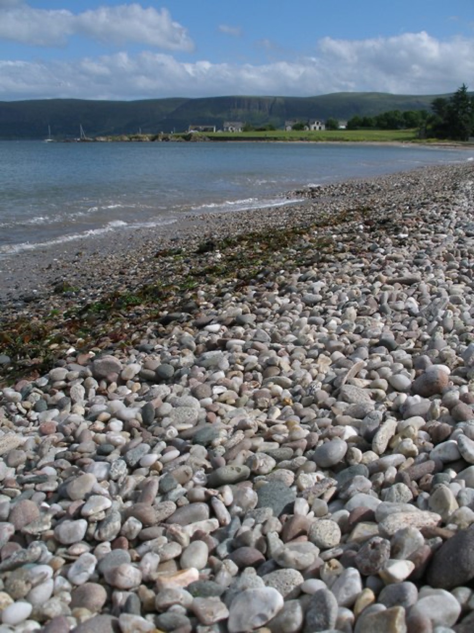 The pebble-and-shingle shore at Cushendall Beach curving away toward the green headland and moored boats under a summer sky