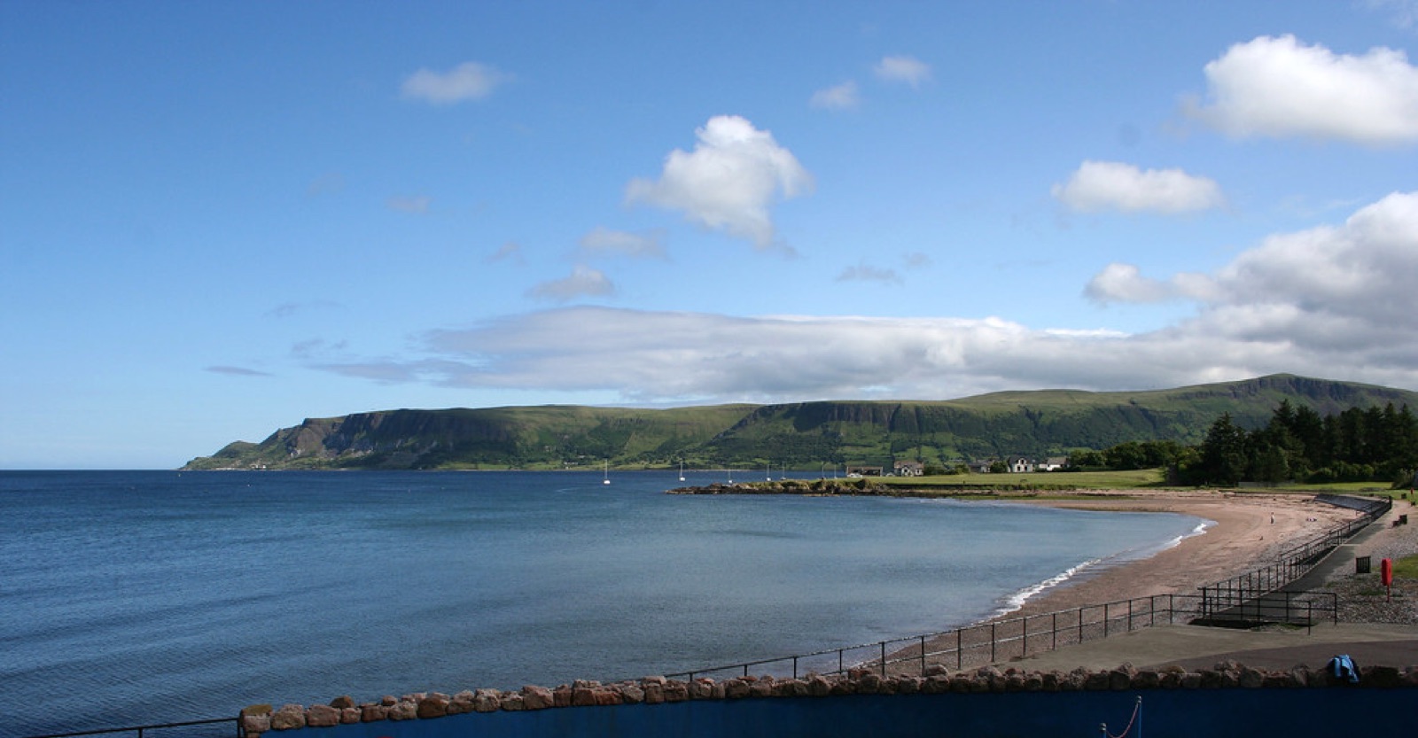 The curving sand-and-shingle beach at Cushendall with a promenade rail, sailing boats moored offshore and the flat-topped Lurigethan headland across Red Bay under a bright blue sky