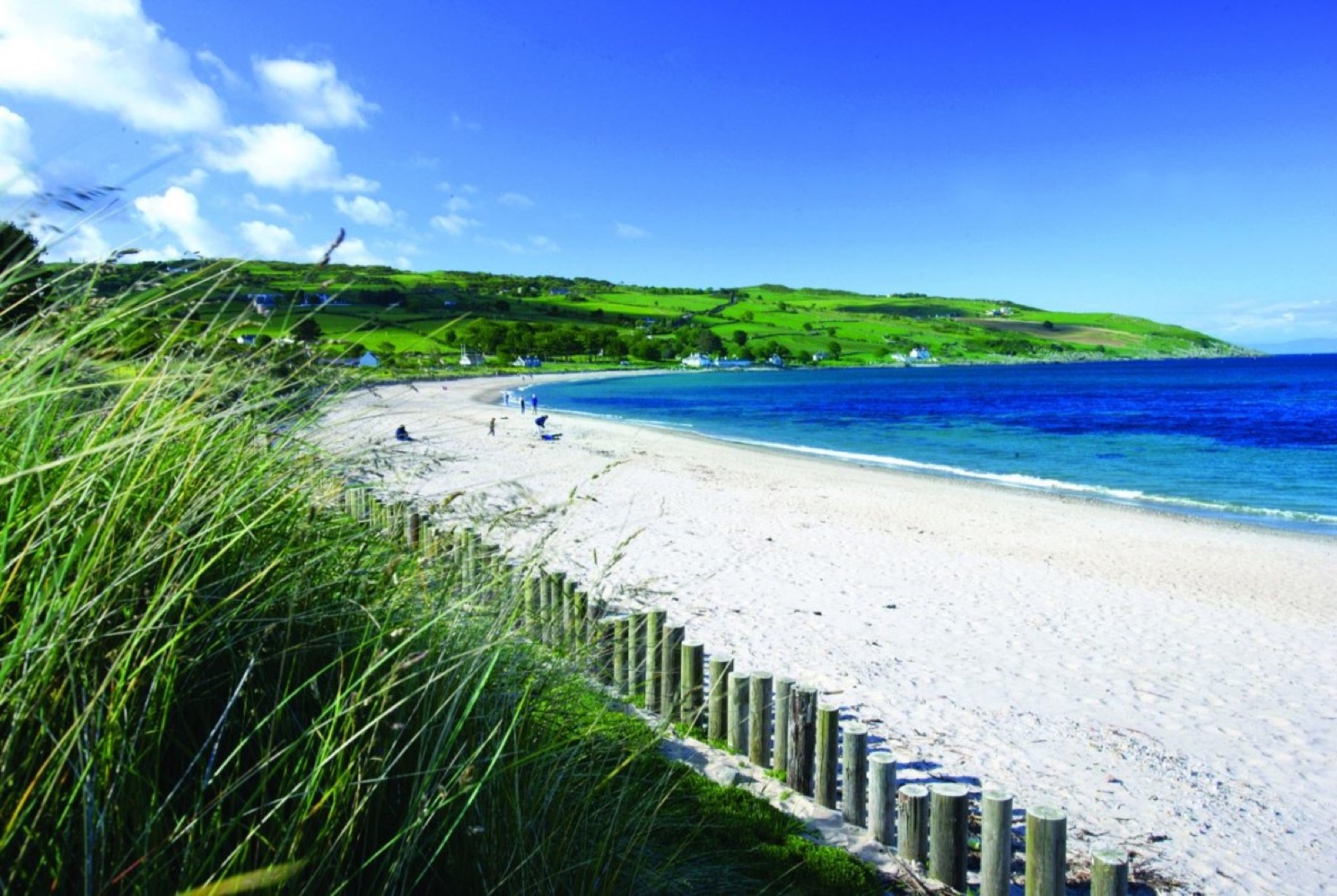 The white sandy beach at Cushendun curving round the bay, with green Glens of Antrim hills behind and marram grass in the foreground under a bright blue sky