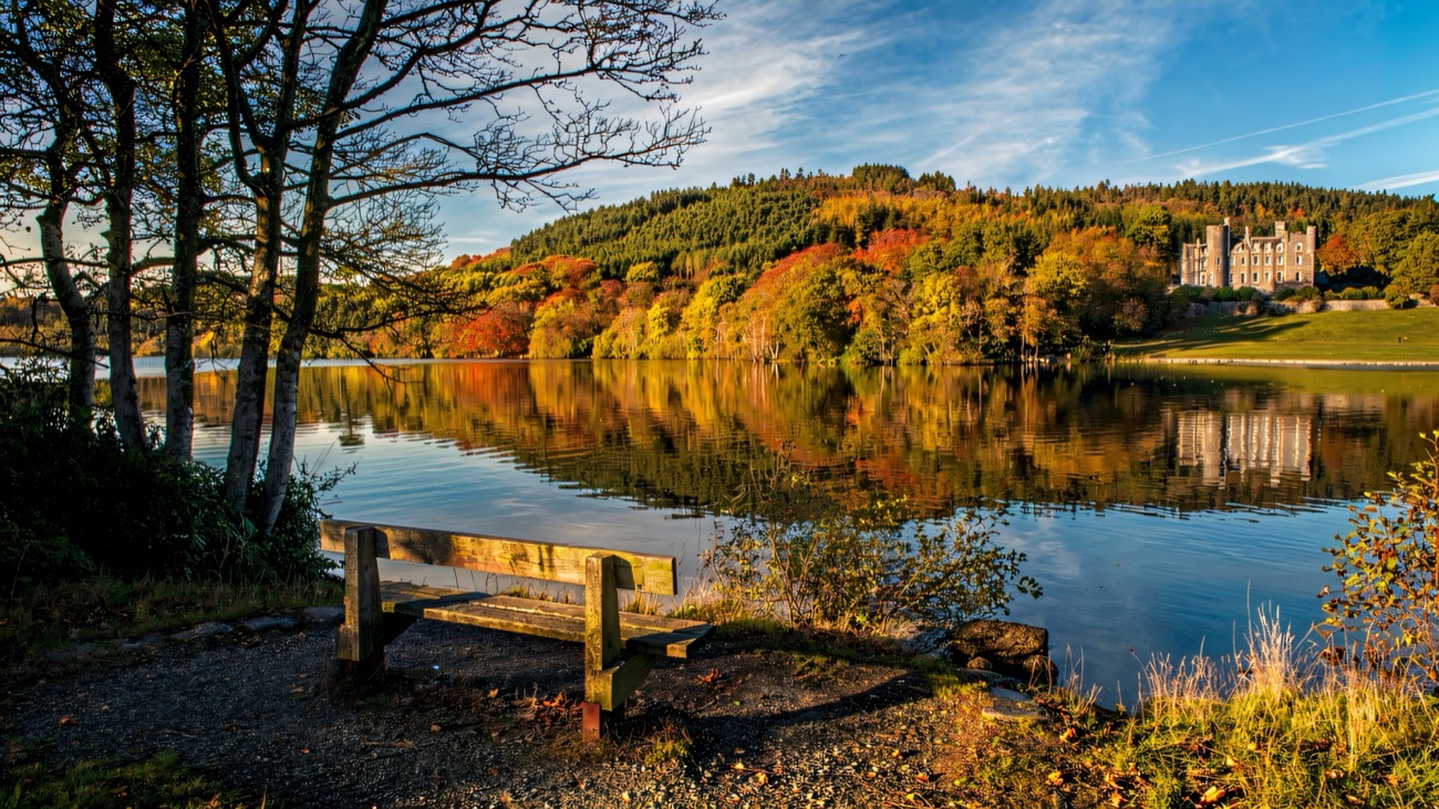 Castlewellan Castle and autumn woodland reflected in the still lake, with a wooden bench in the foreground