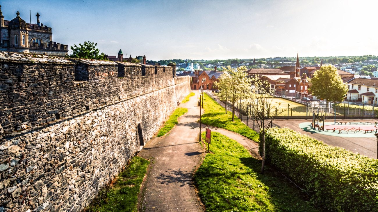 The broad stone wall walkway with a path, green space and the city beyond under blue sky