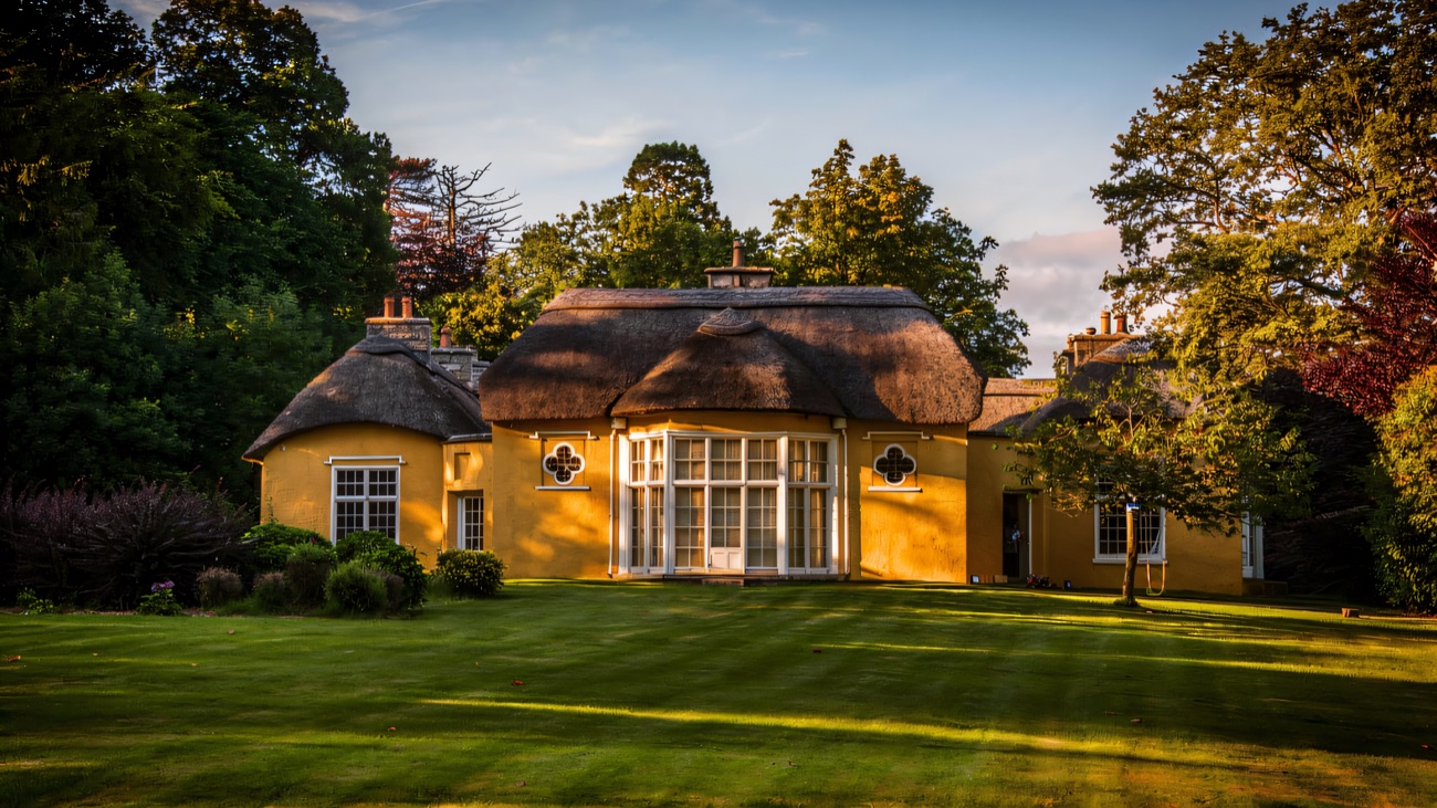 Derrymore House lit by golden evening light, the central bow window and thatched roof framed by tall trees and open lawn