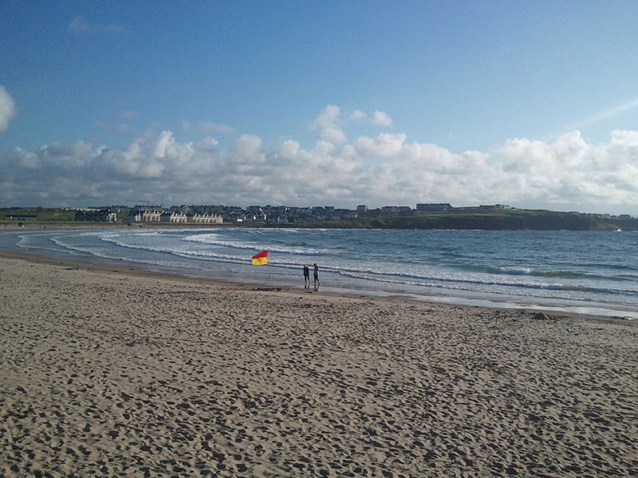 The wide sands of East Strand with a red-and-yellow lifeguard flag and the town of Portrush across the bay