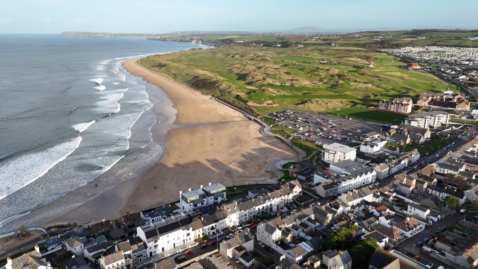Aerial view of East Strand curving below the Royal Portrush golf links, with the town in the foreground and surf rolling in