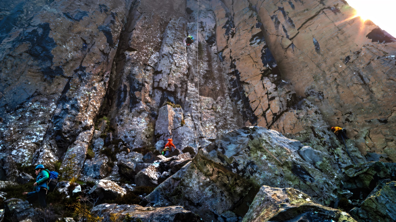 The wide crag face, with tiny roped climbers above the boulder field