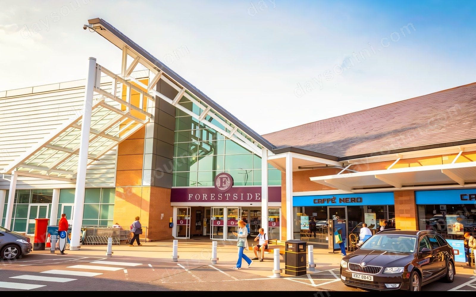 The main entrance at Forestside Shopping Centre in the evening sun, with Caffè Nero beside the doors and shoppers coming and going