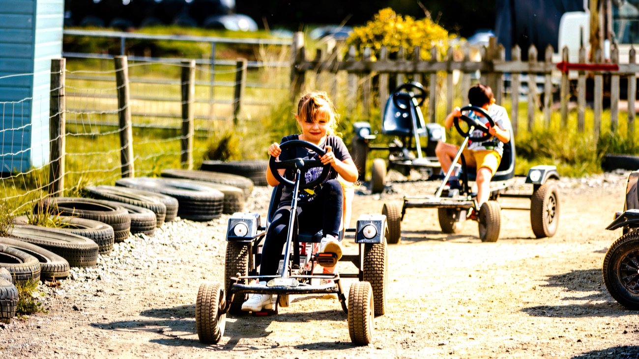 A young child pedalling a go-kart along a gravel track lined with tyres at a family farm