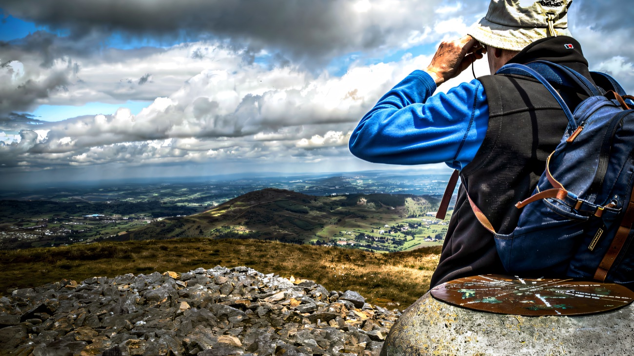 A walker with binoculars beside a summit cairn, valley stretching below