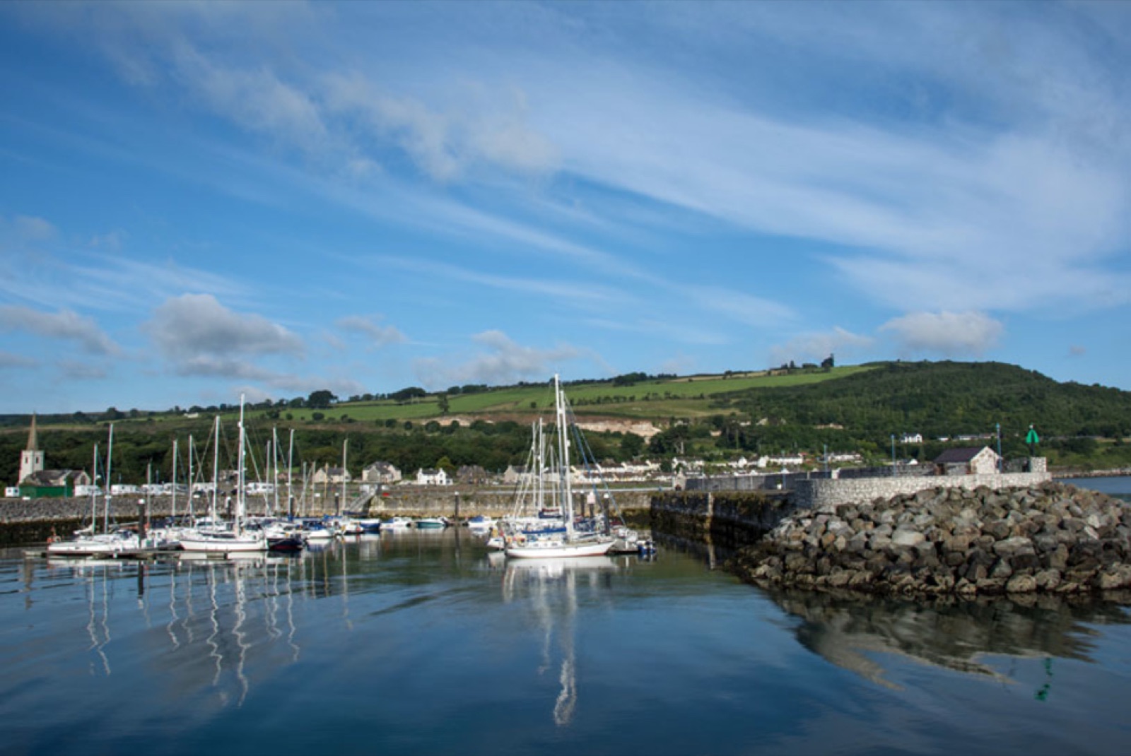 Yachts moored in Glenarm's limestone marina with reflections in still water, the village and the green Glens hillside behind under a bright blue sky