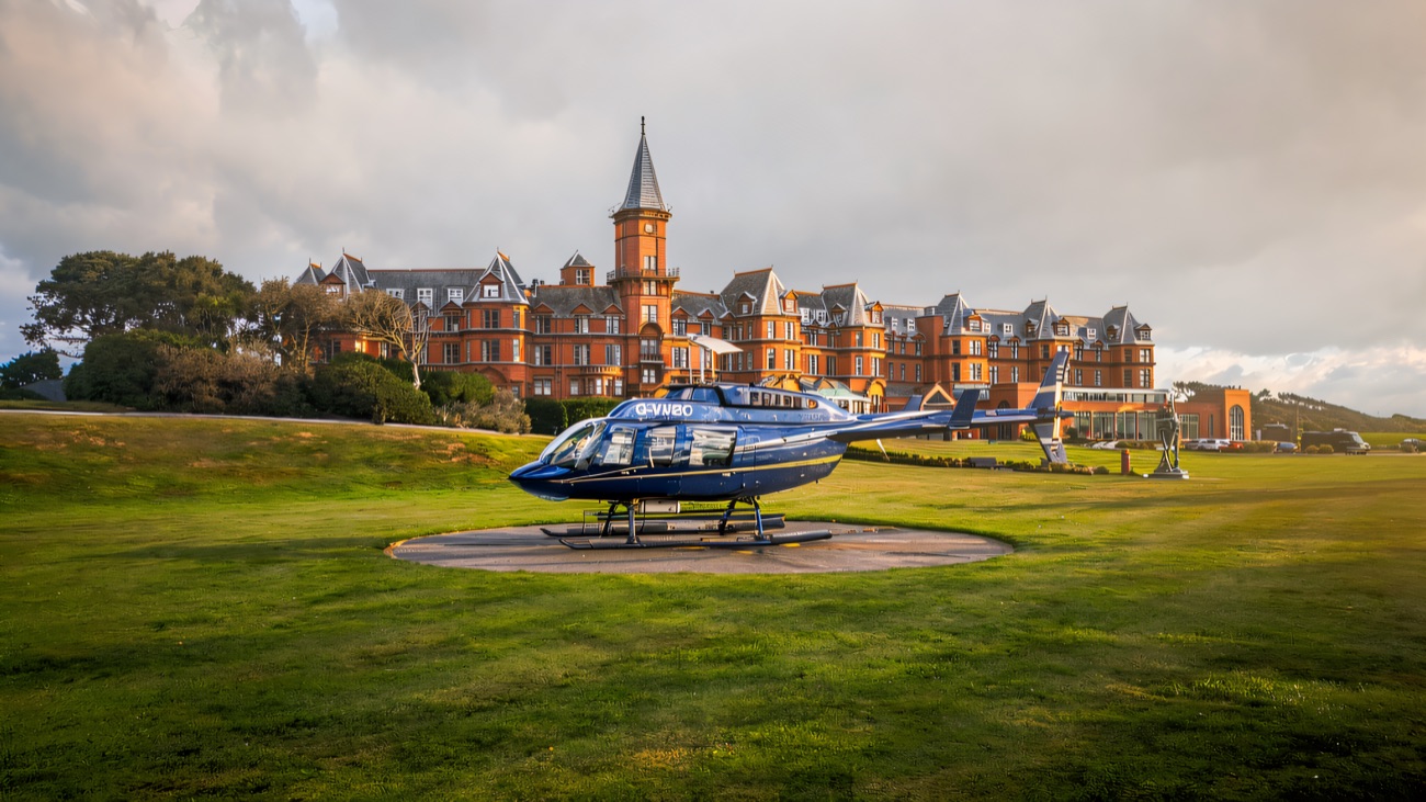 A blue helicopter resting on a circular landing pad on a wide lawn, with a grand red-brick hotel behind
