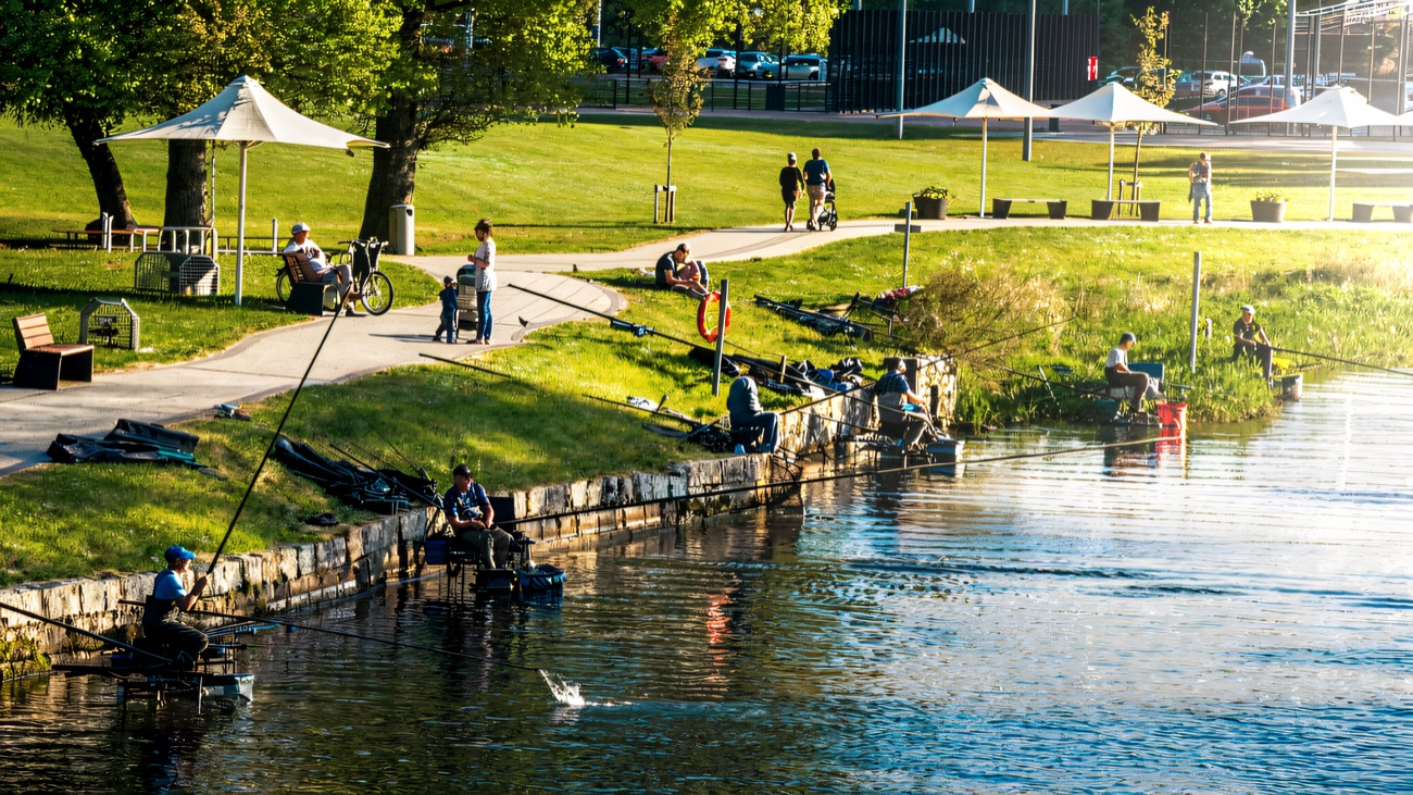 Anglers spaced along the stone-edged coarse fishing stands on the water's edge, with parkland and shade sails behind