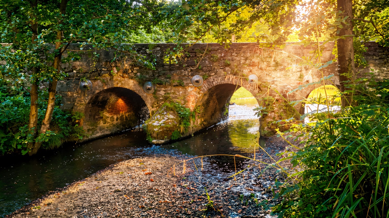 A weathered stone arch bridge crosses the water amid summer green