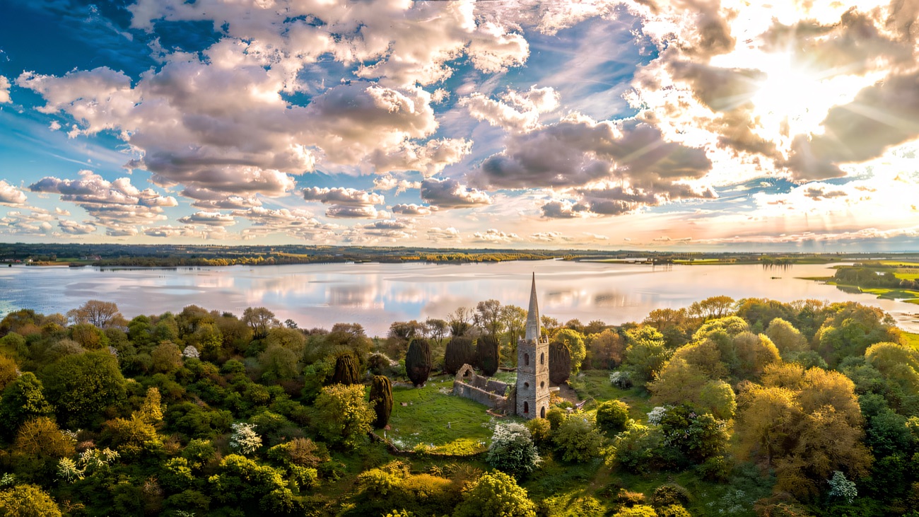 An aerial view of Church Island's slender spire rising from blossom-flecked woodland, the lough stretching behind.
