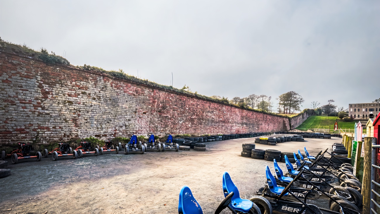 A row of pedal go-karts on the gravel track beside the tall old brick wall