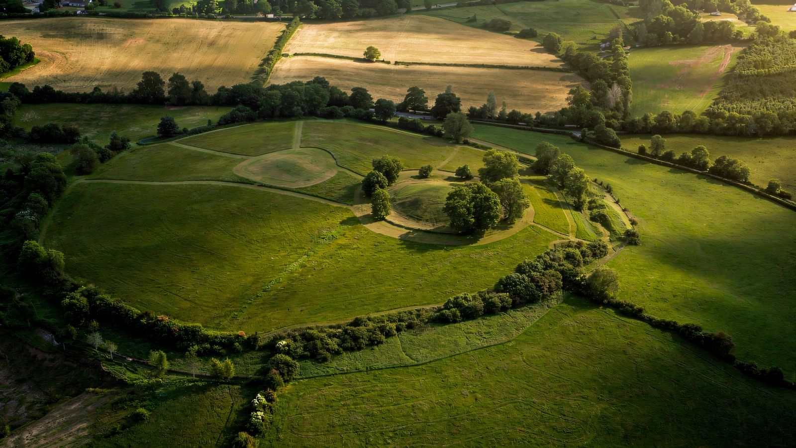 Aerial view of the great circular earthwork of Navan Fort, its central grassy mound ringed by a bank and ditch, surrounded by farmland