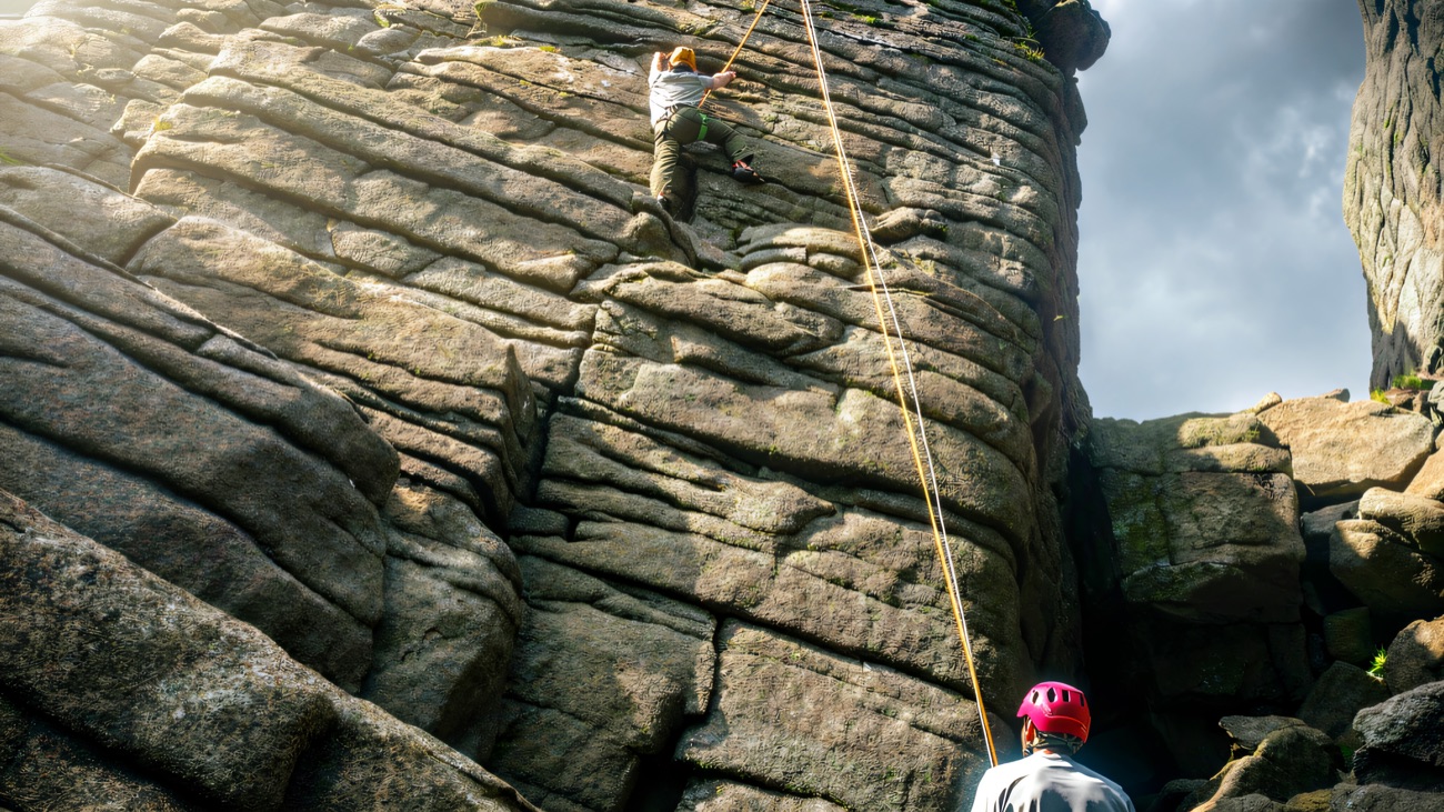 A climber working up a cracked granite face on a rope, with a belayer waiting at the foot of the crag