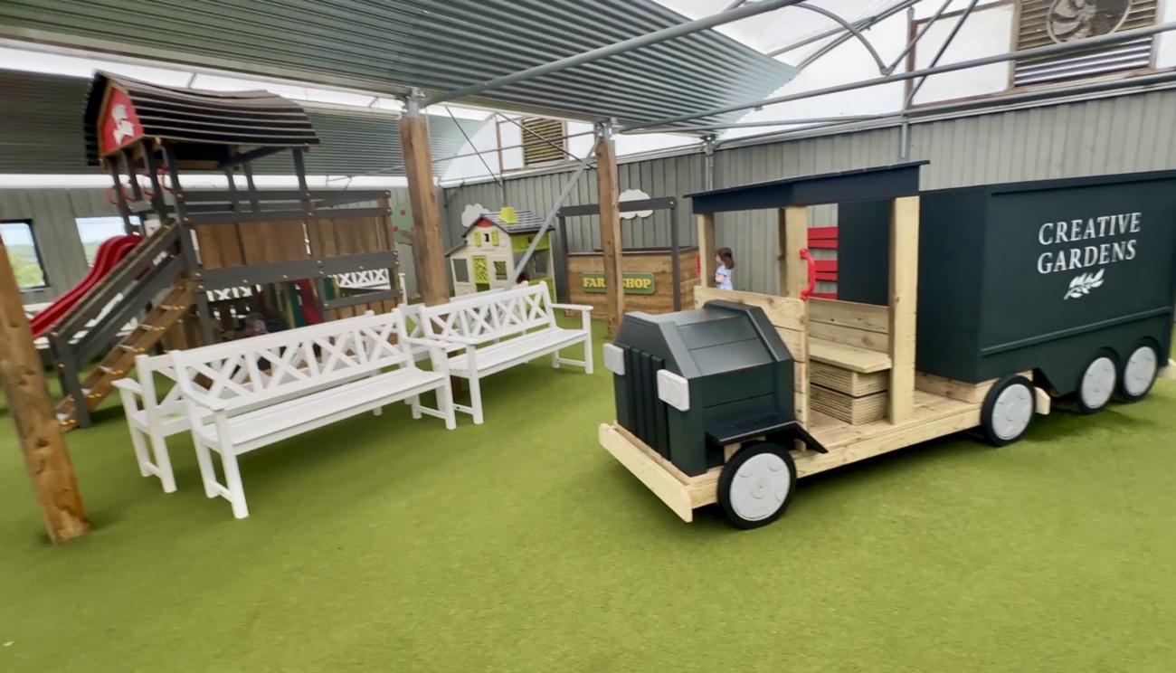 A large wooden 'Creative Gardens' delivery-truck play structure and a climbing frame with a red slide, on astroturf under the canopy