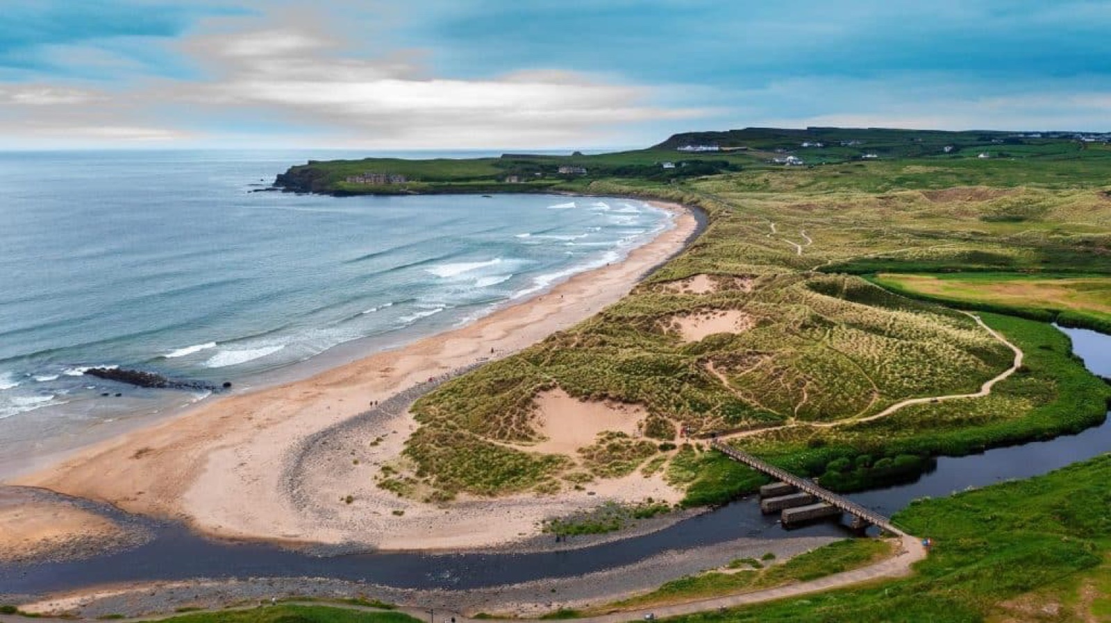Bushfoot Strand curving round to the mouth of the River Bush at Portballintrae, with grassy dunes, a footbridge and the Causeway Coast headland beyond