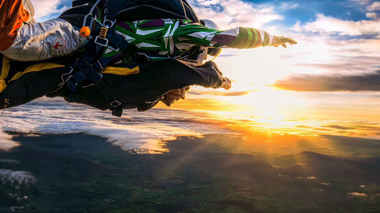 Skydivers in freefall high above green fields, with the low sun lighting the cloud tops