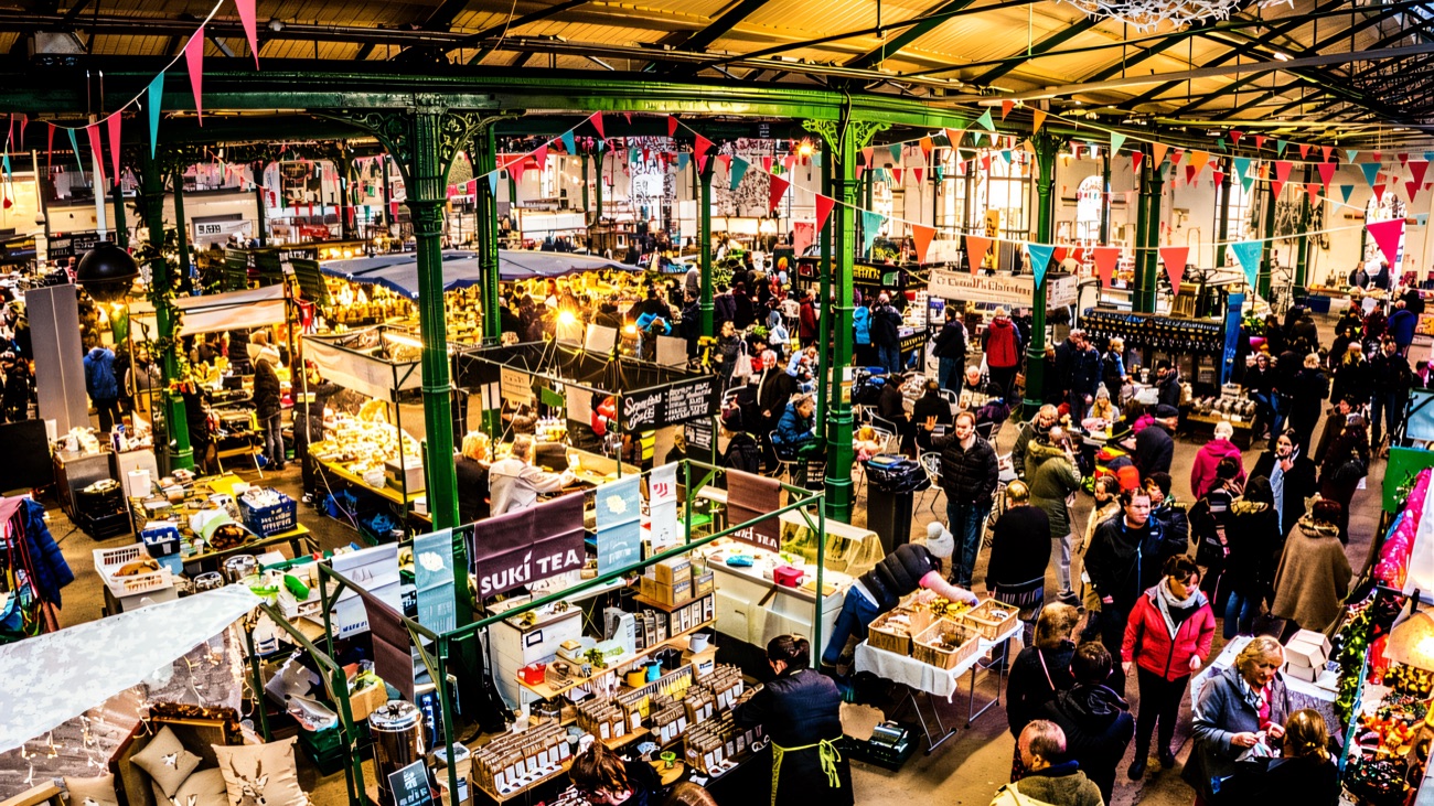 Looking down over the busy market hall — rows of food and craft stalls, bunting overhead and crowds browsing between them