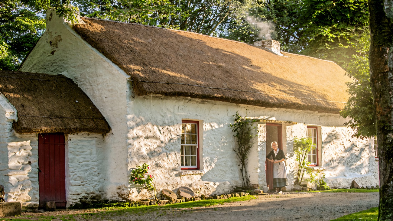 A whitewashed thatched Ulster cottage with red door and window frames, a costumed figure in the doorway