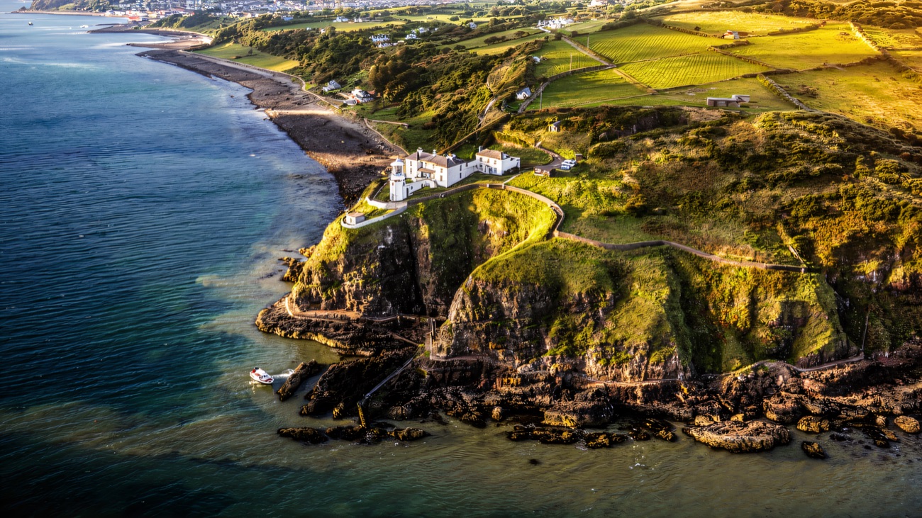 Blackhead lighthouse standing white on the clifftop above clear green water, Whitehead in the distance